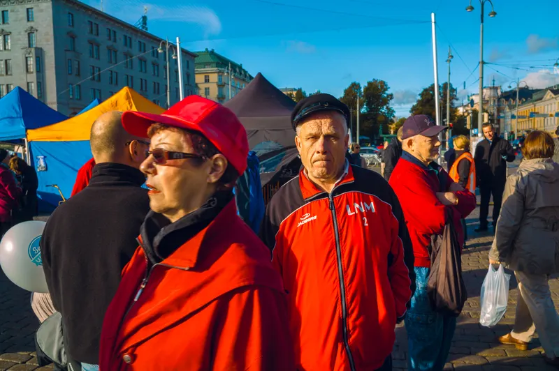 A woman in a red cap and sunglasses stands amid a crowd in red jackets at an outdoor market with colorful tents and a balloon.