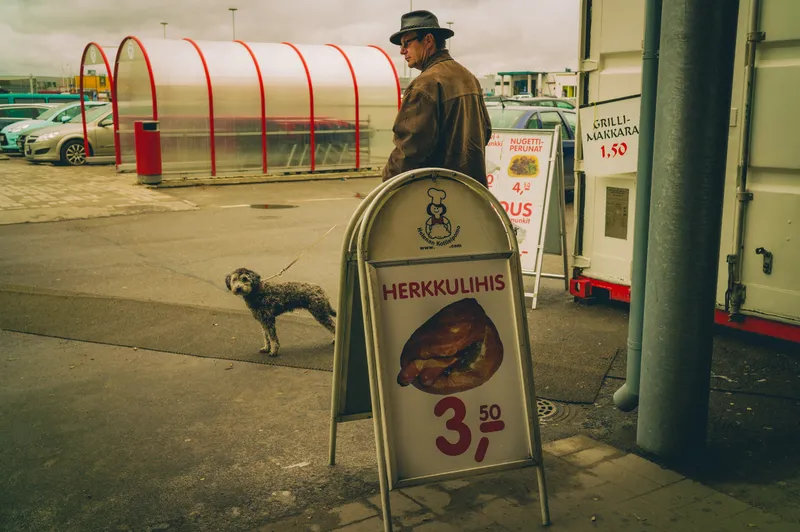 A man in a hat walks a shaggy dog past a food-stall sandwich board advertising snacks in a parking lot market.