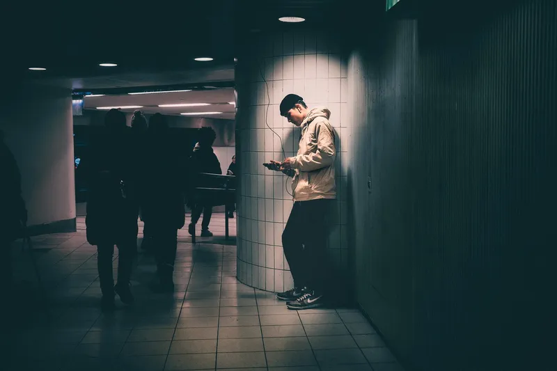 A man leans against a tiled pillar checking his phone in a metro corridor, silhouetted figures passing behind him in the dim light.