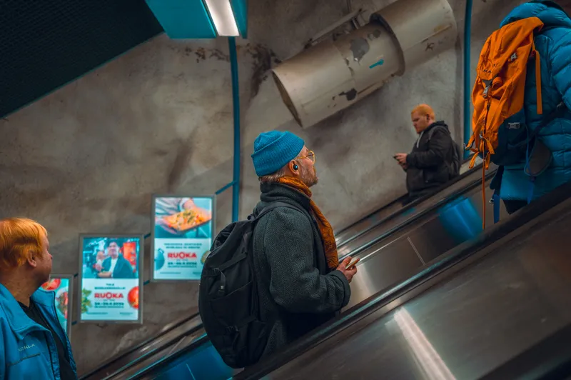 A man in a blue beanie and scarf rides a metro escalator past illuminated ads, a hiking backpack hanging on the rail beside him.