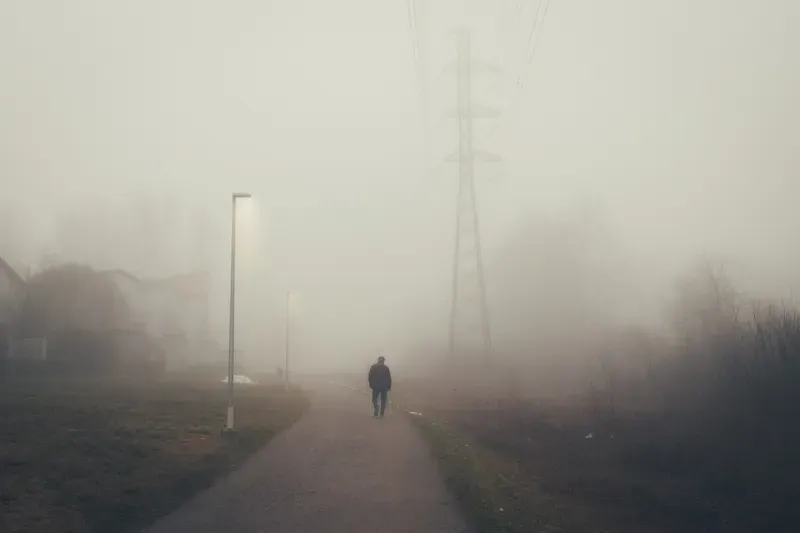 A man using a cane walks away down a misty path past houses and a power line pylon.