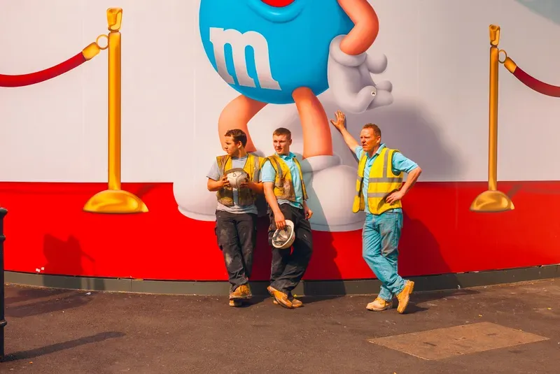 Three construction workers in hi-vis vests lean against a giant M&M's World mural, one resting a hand on the character's painted hand.