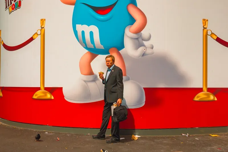A man in a suit eats a sandwich while walking past the M&M's World mural, a pigeon pecking at litter nearby.