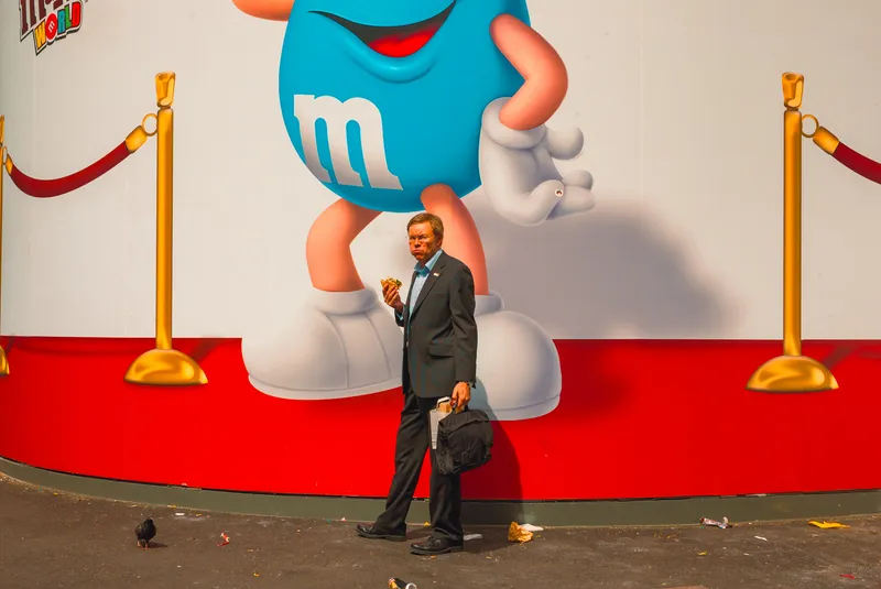 A man in a suit eats a sandwich while walking past the M&M's World mural, a pigeon pecking at litter nearby.