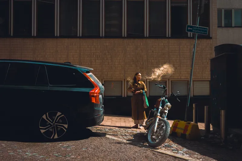 A woman in a trench coat exhales smoke in low golden sunlight beside a parked motorcycle and a black SUV.