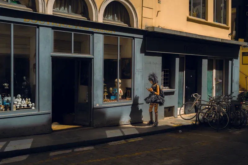 A woman in a black tutu and fishnet tights checks her phone outside an antique shop whose window is crowded with busts and figurines.