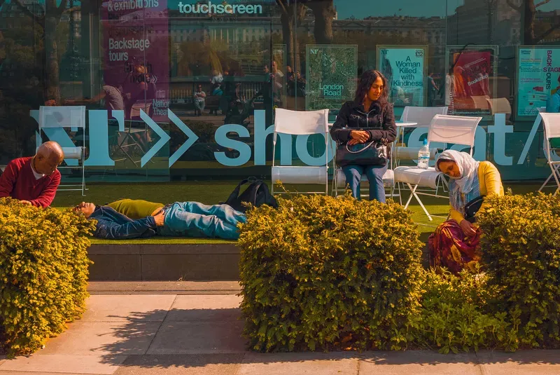 Visitors relax and lie on artificial turf in front of the National Theatre's glass signage reading "shop", posters visible behind them.