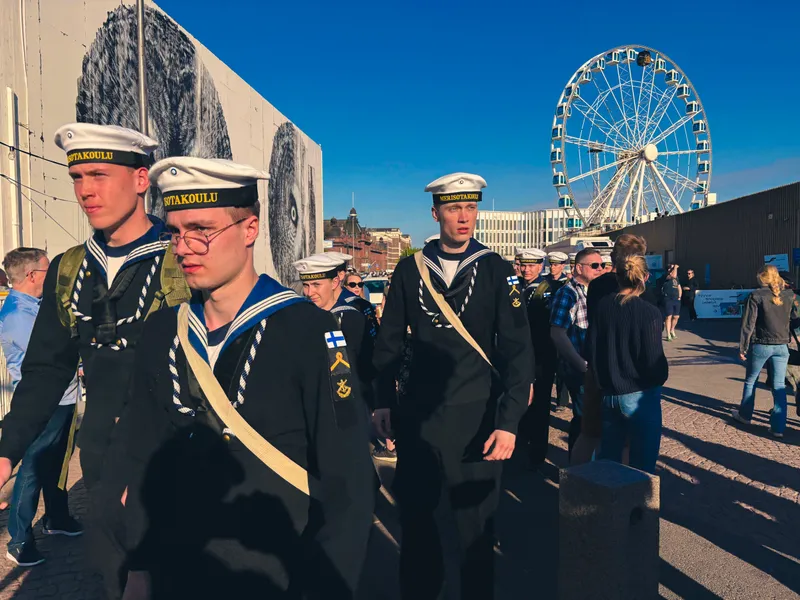 Young naval cadets in sailor uniforms and white caps walk past a mural under a bright blue sky, a ferris wheel turning behind them.