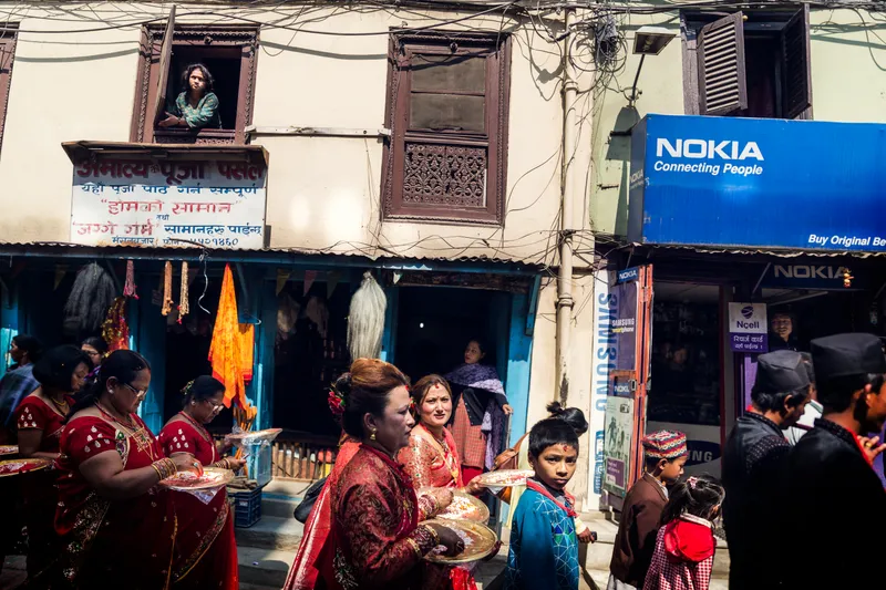 Women in red saris carry ceremonial plates through a busy street lined with shopfronts, a curious face watching from a window above.