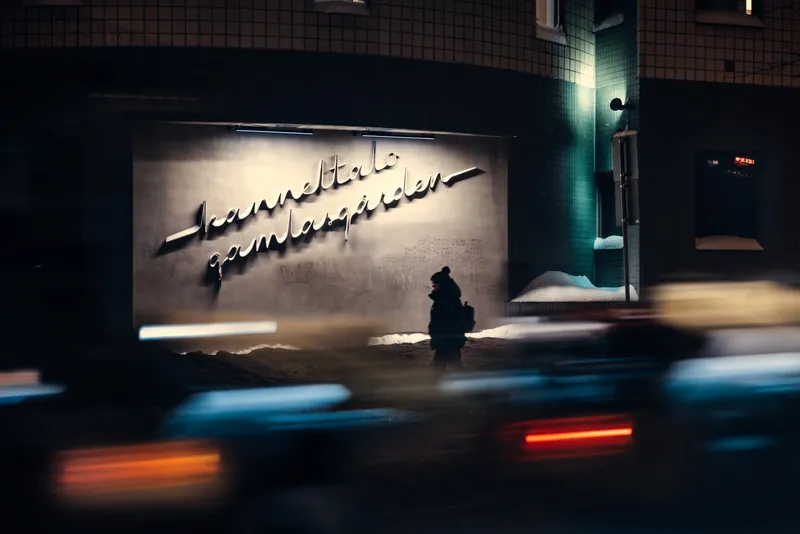A silhouetted figure stands beneath an illuminated cursive sign reading Kanneltalo Gamlagården at night, streaks of car light blurring past in the foreground.