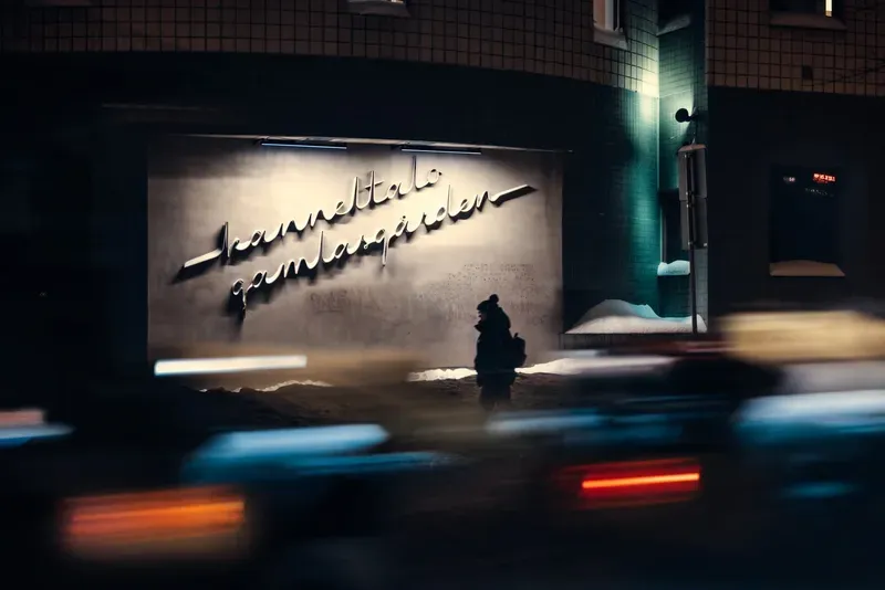 A silhouetted figure stands beneath an illuminated cursive sign reading Kanneltalo Gamlagården at night, streaks of car light blurring past in the foreground.