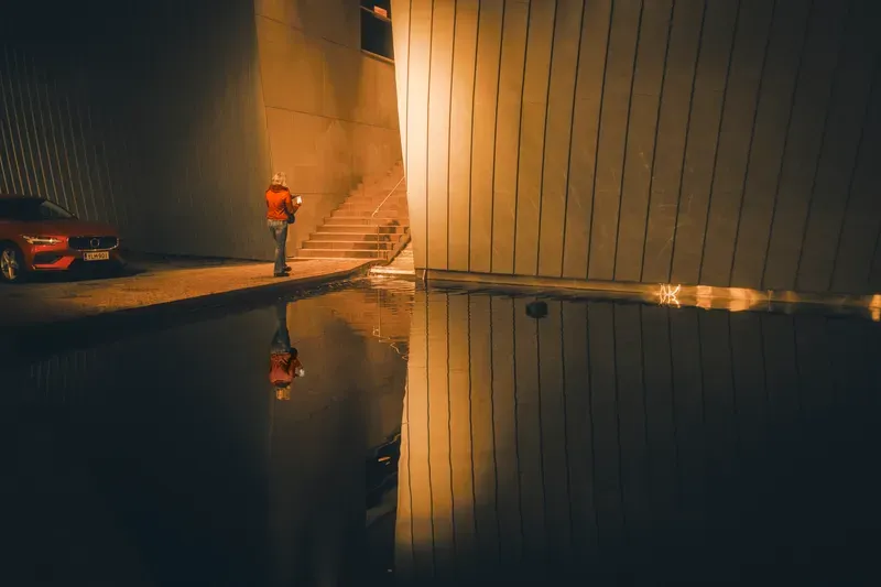 A person in an orange top checks their phone at the foot of a staircase between two curved buildings at night, their reflection and a parked red car mirrored in a puddle below.