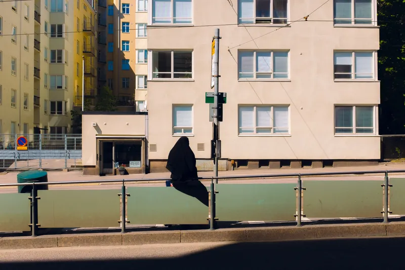 A woman in a black niqab sits on a railing across from a sunlit apartment building, sharp shadows cutting across the street.