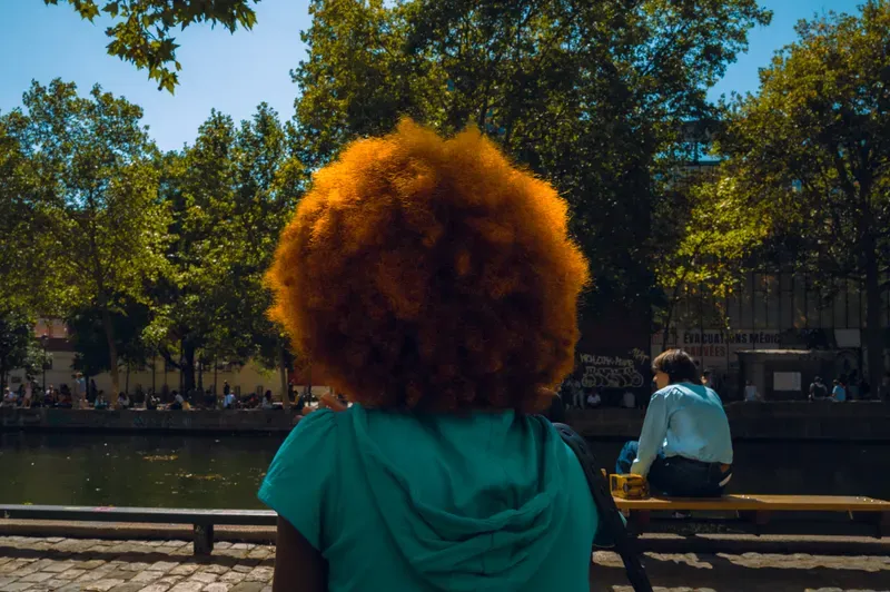 Seen from behind, a person with a large orange afro sits facing a tree-lined canal, another figure resting on a bench beyond.