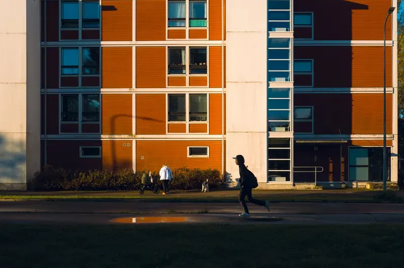 A figure in a black hoodie and cap runs along a path in low golden light past an orange apartment block, a woman with a pram and a small dog by the hedge behind.