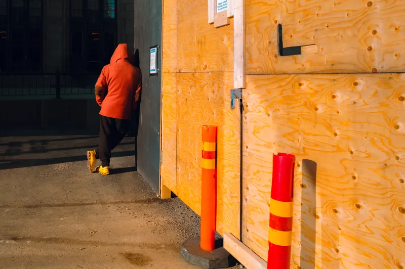 A hooded figure in orange leans against a gap in a plywood construction hoarding, warm evening light raking across the wood grain.