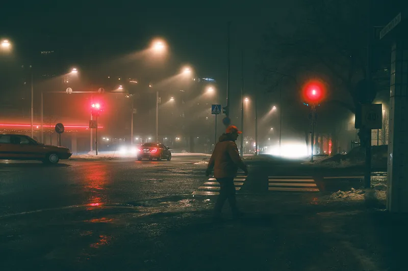 A man in an orange jacket and cap crosses a wet city intersection beneath a red traffic light, car headlights glowing through the night.