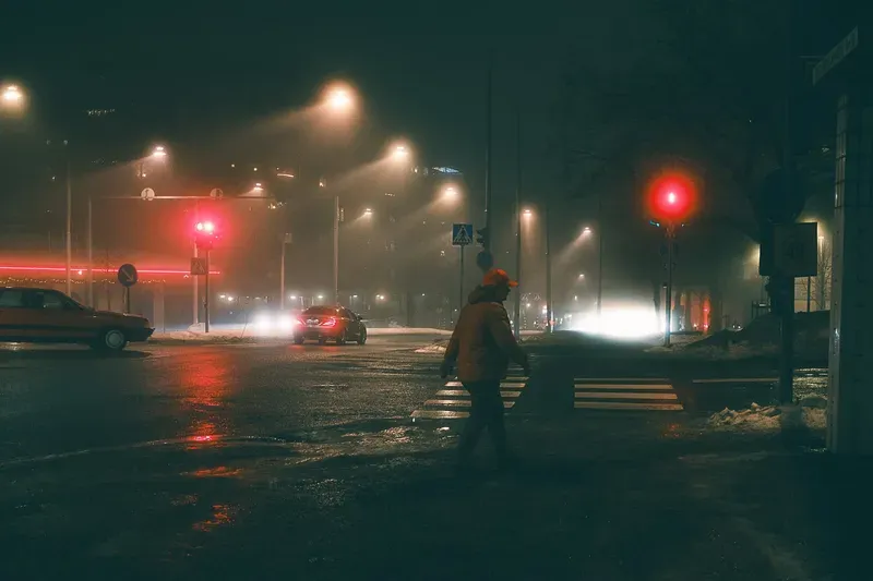 A man in an orange jacket and cap crosses a wet city intersection beneath a red traffic light, car headlights glowing through the night.