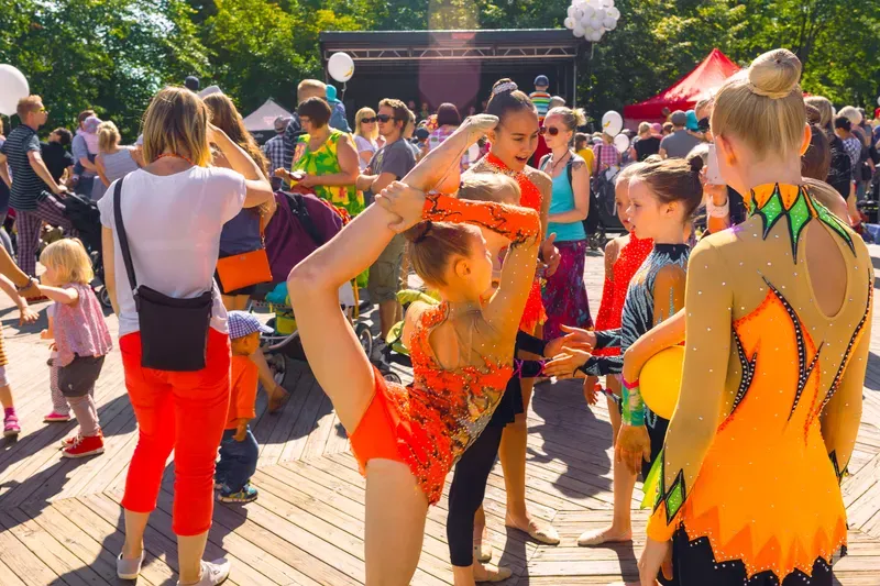 Young rhythmic gymnasts in orange and gold leotards stretch and chat in front of a crowd at an outdoor stage, one girl holding her leg up beside her head.