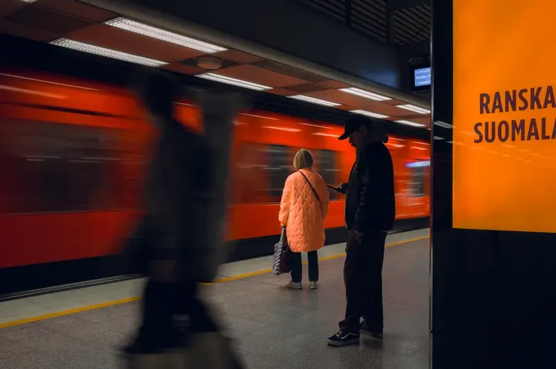 A woman in a quilted peach coat and a man in a cap checking his phone wait on a metro platform as a red train streaks past, a blurred walker in the foreground and a glowing orange advertising panel filling the right edge.