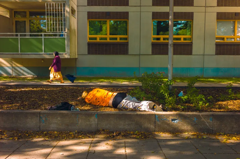 A woman in a headscarf walks past a person lying face-down in a mulch flowerbed in front of an apartment building.