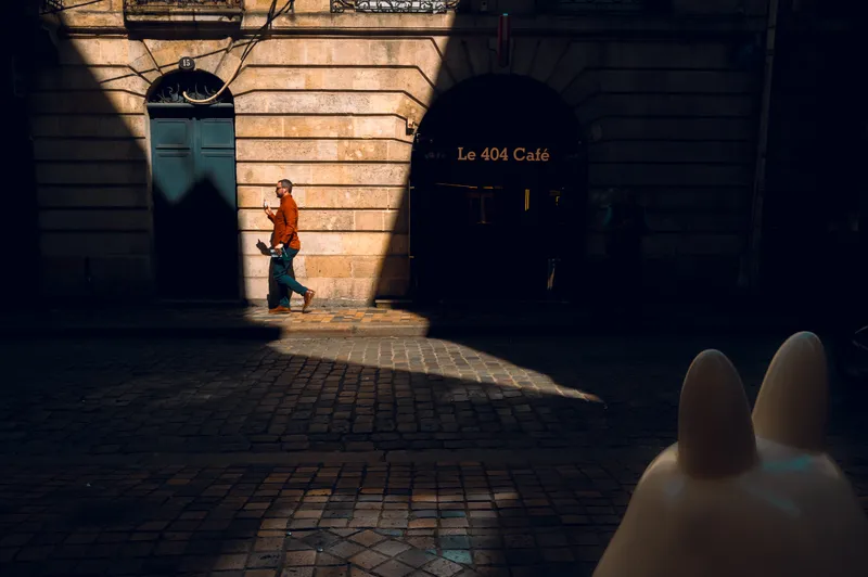 A man in an orange shirt checks his phone in a strip of sunlight beside a stone archway leading to Le 404 Café.