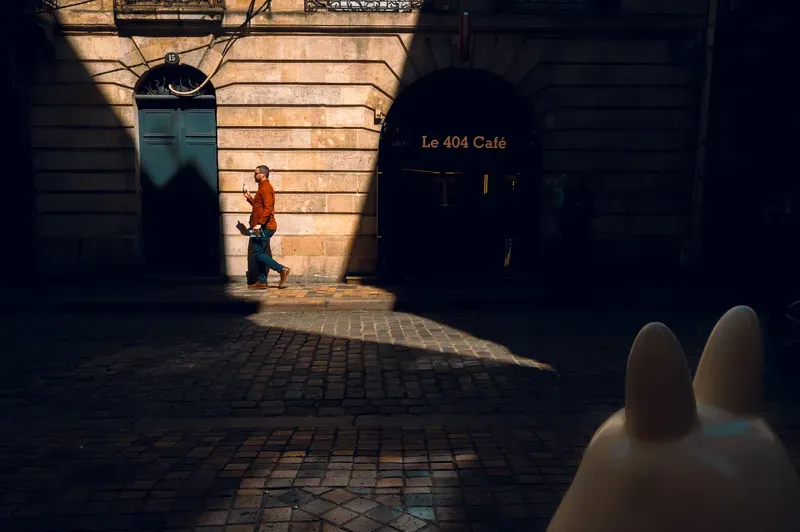 A man in an orange shirt checks his phone in a strip of sunlight beside a stone archway leading to Le 404 Café.
