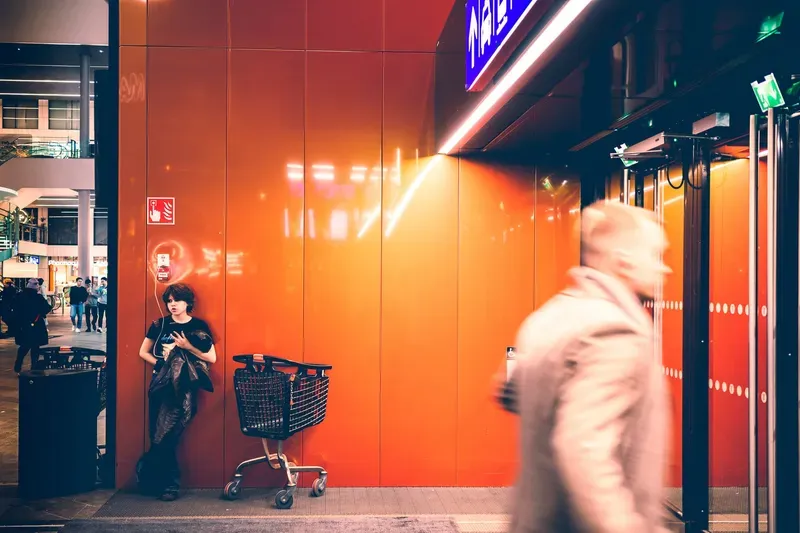 A girl leans against a glowing orange wall beside an empty shopping cart while a blurred figure hurries past in a mall corridor at night.
