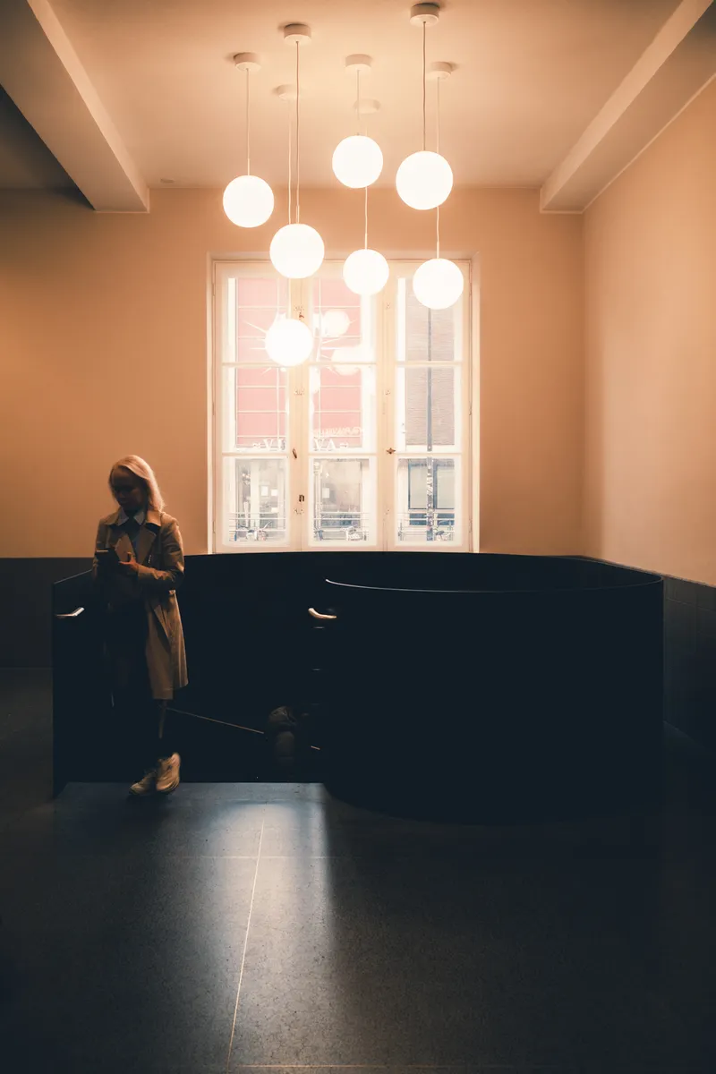 A woman checks her phone beside a dark curved staircase, a cluster of round pendant lights glowing above her in a peach-toned room.