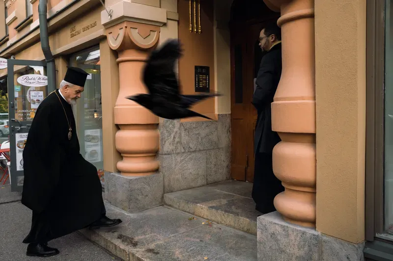 An Orthodox priest in black robes and a tall hat steps up to a doorway between peach-coloured columns as a crow blurs past at head height, a second clergyman waiting in the entrance.