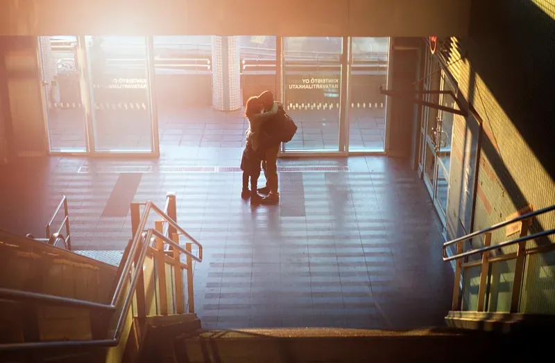 A couple embraces in a sunlit station entryway, warm flare and long shadows streaming across the tiled floor and staircase.