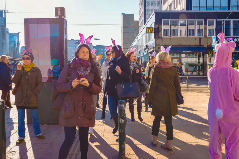Passers-by on a sunlit city street wear pink bunny ears, a person in a full pink bunny costume at the edge of the frame and a McDonald's sign behind them.