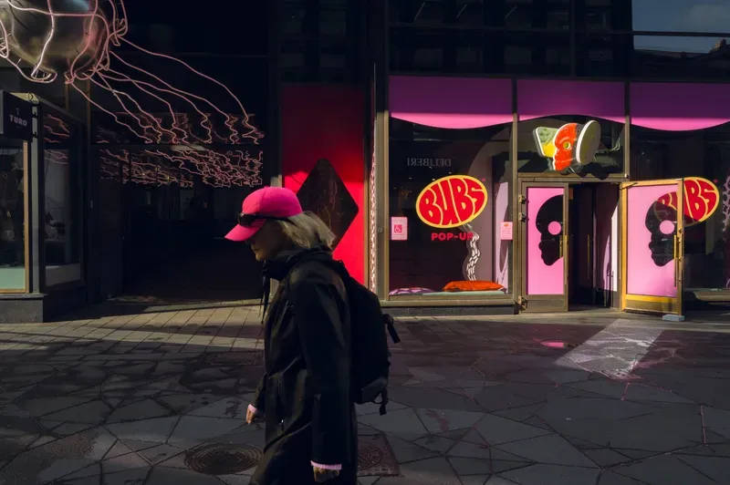 A woman in a hot pink cap and sunglasses walks through shadow past the magenta windows of a sweet shop pop-up, a pink neon jellyfish trailing across the glass behind her.