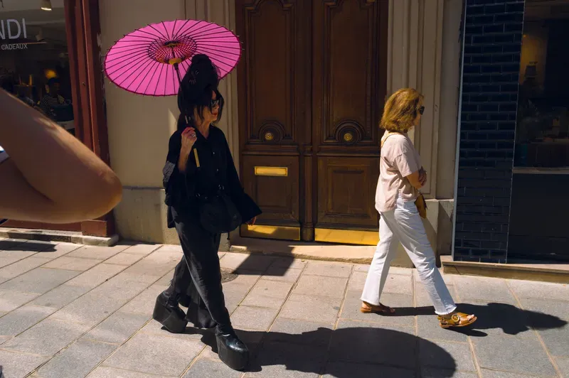 A woman dressed all in black carries a bright pink parasol past a tall wooden door, passing a woman in white walking the other way.