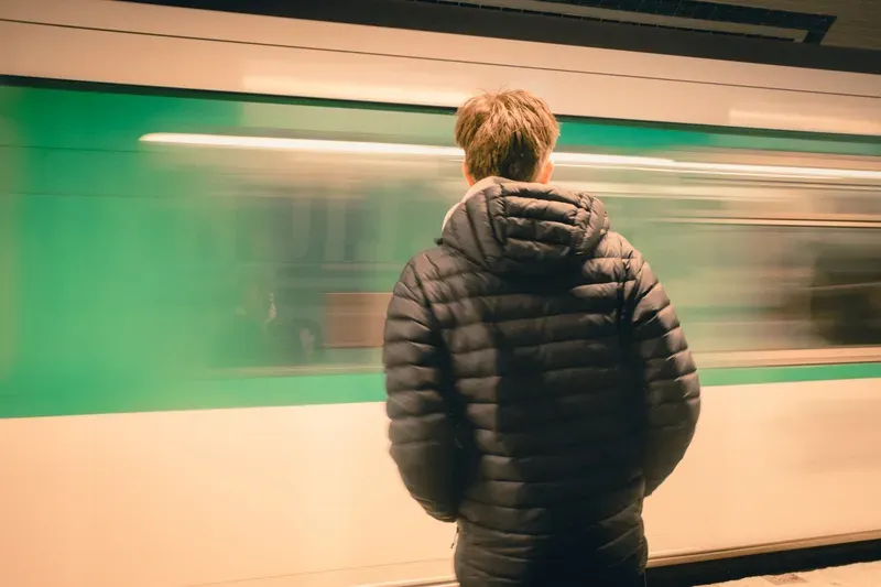 A man in a puffer jacket stands with his back to the camera as a green train blurs past on a station platform.