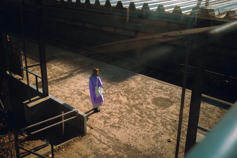 A woman in a purple coat waits alone on an empty train platform, caught in a narrow shaft of light beside the tracks.
