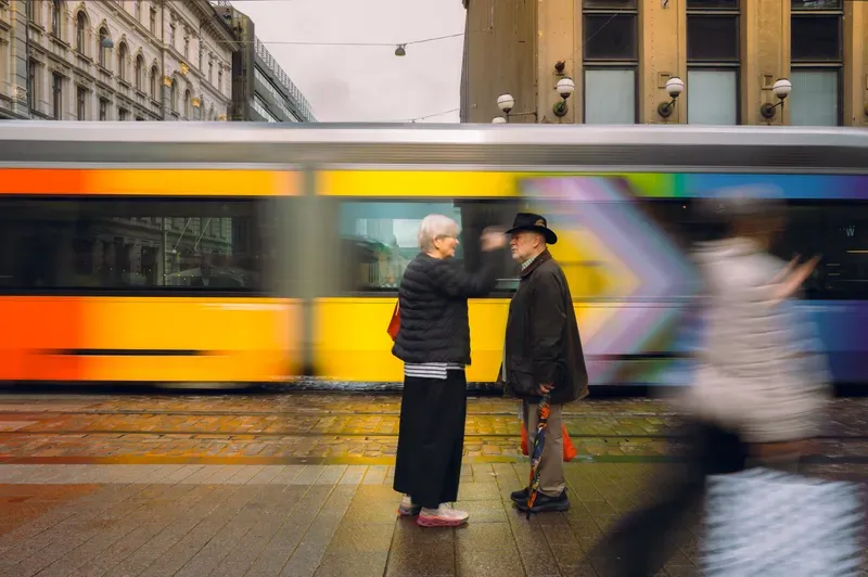 An older woman in a black puffer jacket gestures toward an older man in a wide-brimmed hat holding a folded umbrella as they talk on a wet cobbled street, a blurred yellow and rainbow-striped tram sliding past behind them and a figure smeared by motion in the foreground.
