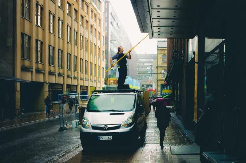 A man stands atop a van reaching a long yellow pole up to clean an awning in a rainy city alley as a woman with an umbrella passes.