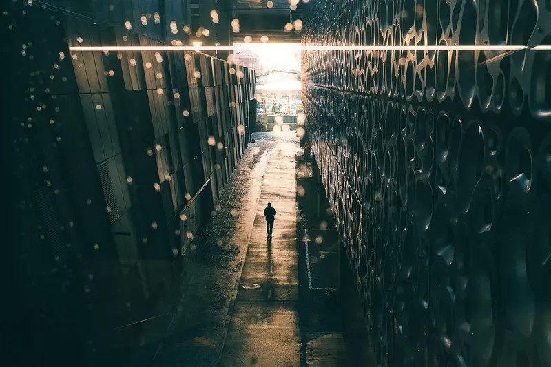 A figure walks toward a bright opening at the end of a narrow alley between a patterned metal facade and a glass building, rain-speckled bokeh scattered across the foreground.