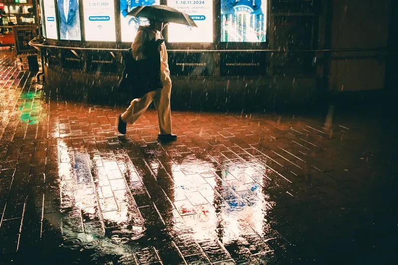 A figure crosses a rain-soaked plaza under a black umbrella, illuminated billboards reflecting in the wet pavement at night.