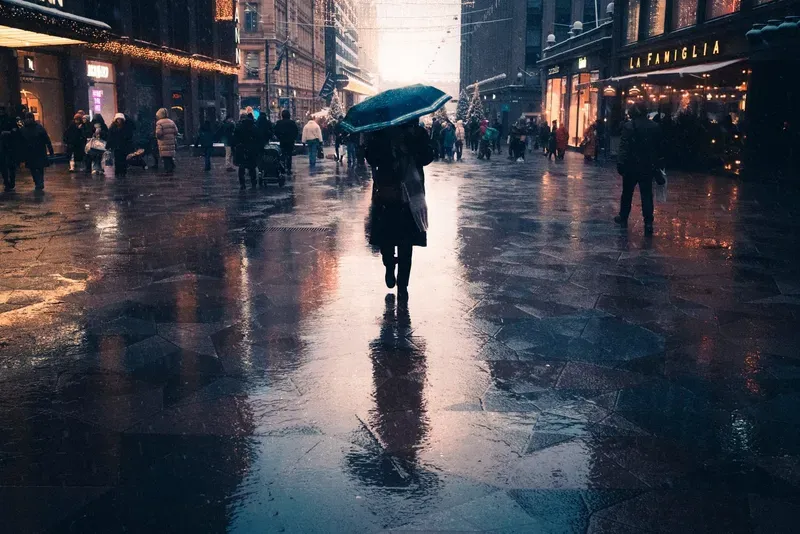 A woman with an umbrella crosses a rain-slicked shopping street reflecting the lights above.