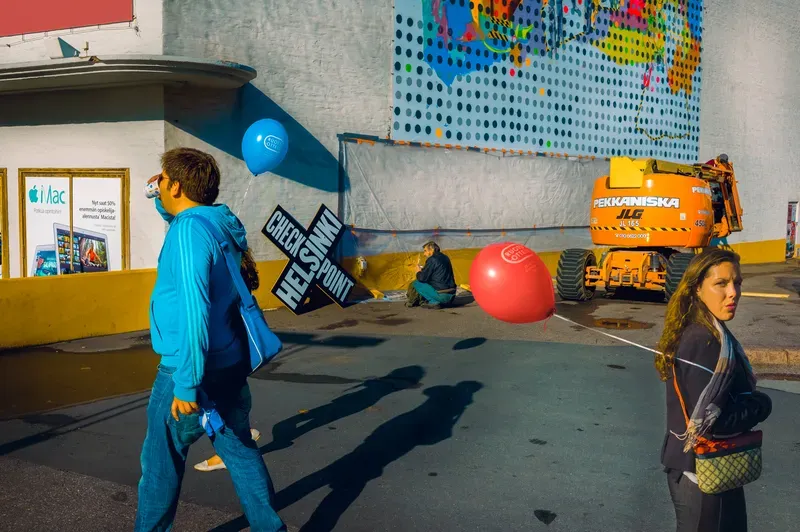 A woman with a red balloon tied to her bag looks back over her shoulder in hard sun while a man in a turquoise hoodie drinks as he walks past, a crossed black sign and an orange lift behind them.