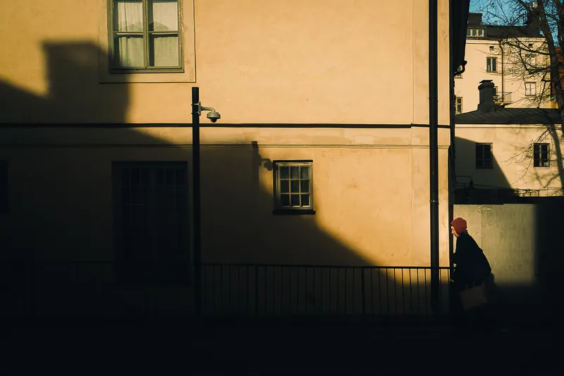 A woman in a red beanie walks past a sunlit apartment facade with a security camera, deep shadows cutting diagonally across the wall.