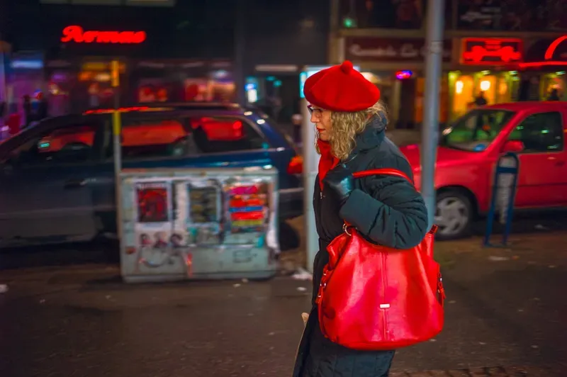 A woman in a red beret and red handbag walks past parked cars at night, neon shop signs glowing behind her.
