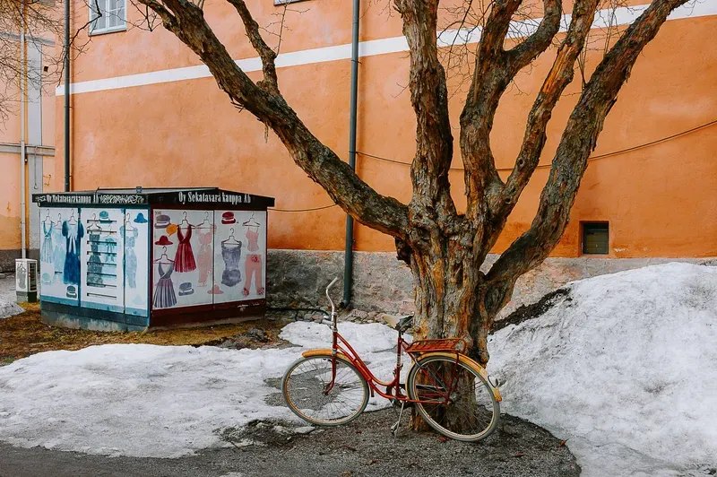 A red bicycle leans against a bare tree trunk beside patches of melting snow and a painted kiosk, in front of an orange building facade.