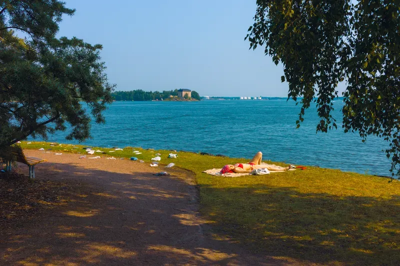 A woman in a red bikini sunbathes on a lakeside lawn beneath overhanging trees, with a small island and ferries visible across the water.