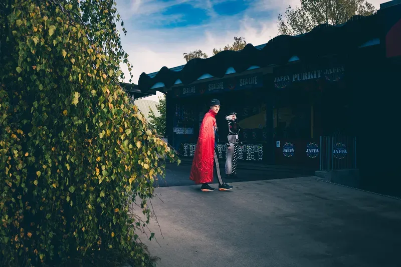 A boy in a red cape glances back while walking past an amusement park kiosk, a costumed performer visible behind him under a blue autumn sky.