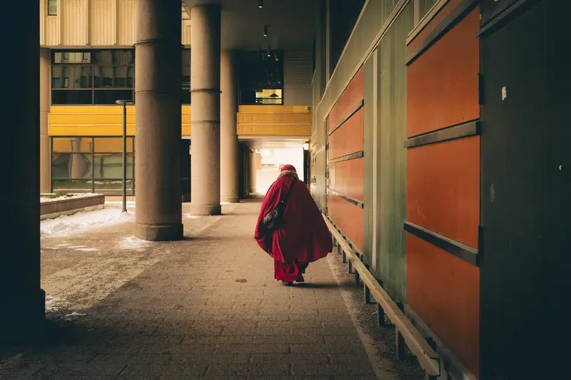 A figure in a flowing red cloak and headscarf walks away down a sunlit concrete corridor between colorful painted panels.