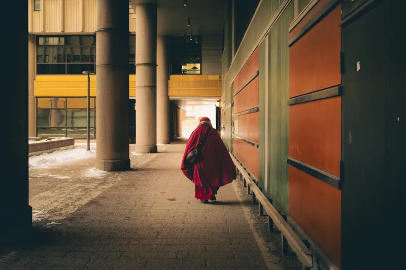 A figure in a flowing red cloak and headscarf walks away down a sunlit concrete corridor between colorful painted panels.