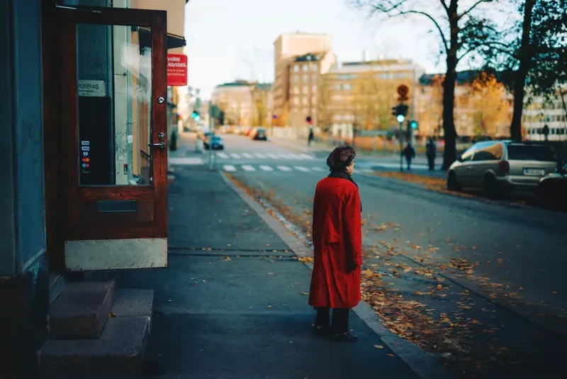 A woman in a red coat and hat waits at a crosswalk on an autumn street, fallen leaves scattered across the pavement.